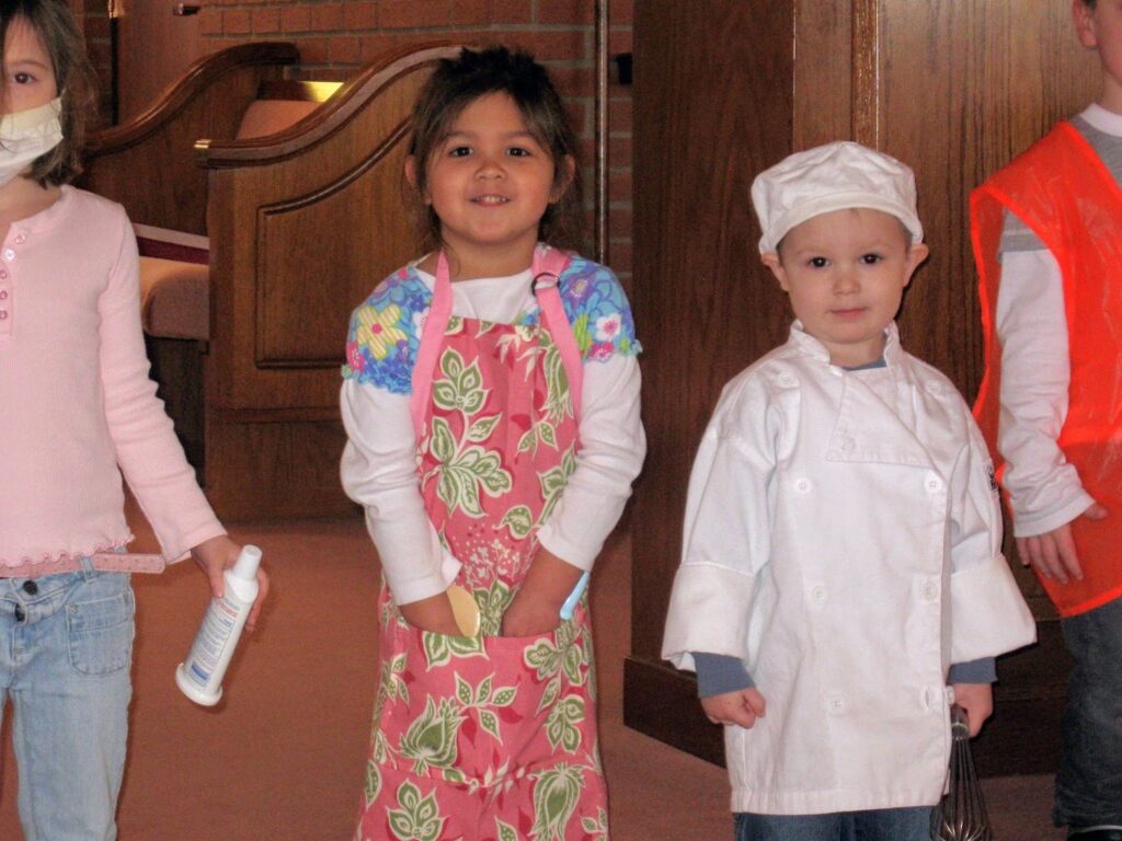 Child learning to bake and is sporting a pink and green apron. Next to her is a child dressed in their chef outfit.