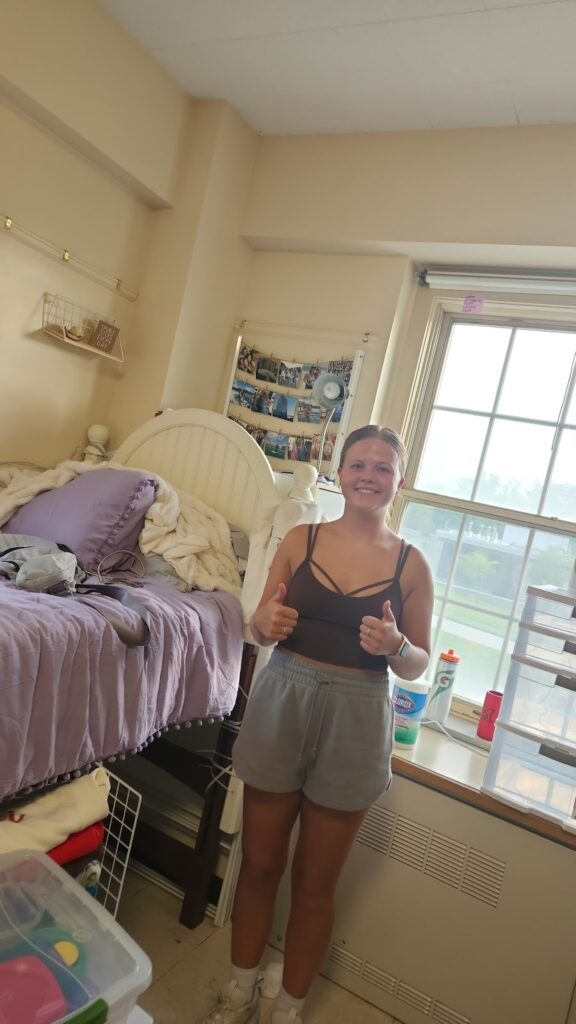 Teen girl standing in a dorm room by a window, giving thumbs up beside a lavender bed with a purple pillow and cozy blanket.