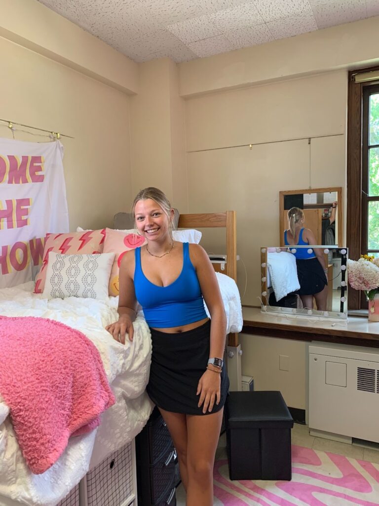 Young woman in a blue top and black skirt standing beside a bed in a pink-themed dorm room, smiling at camera.