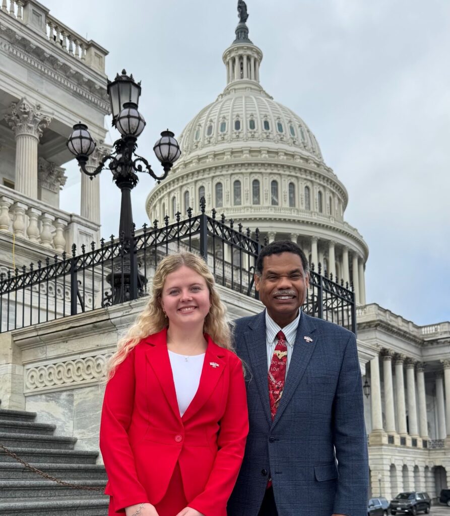 Two people in business attire pose on the steps of the U.S. Capitol with the dome in the background.