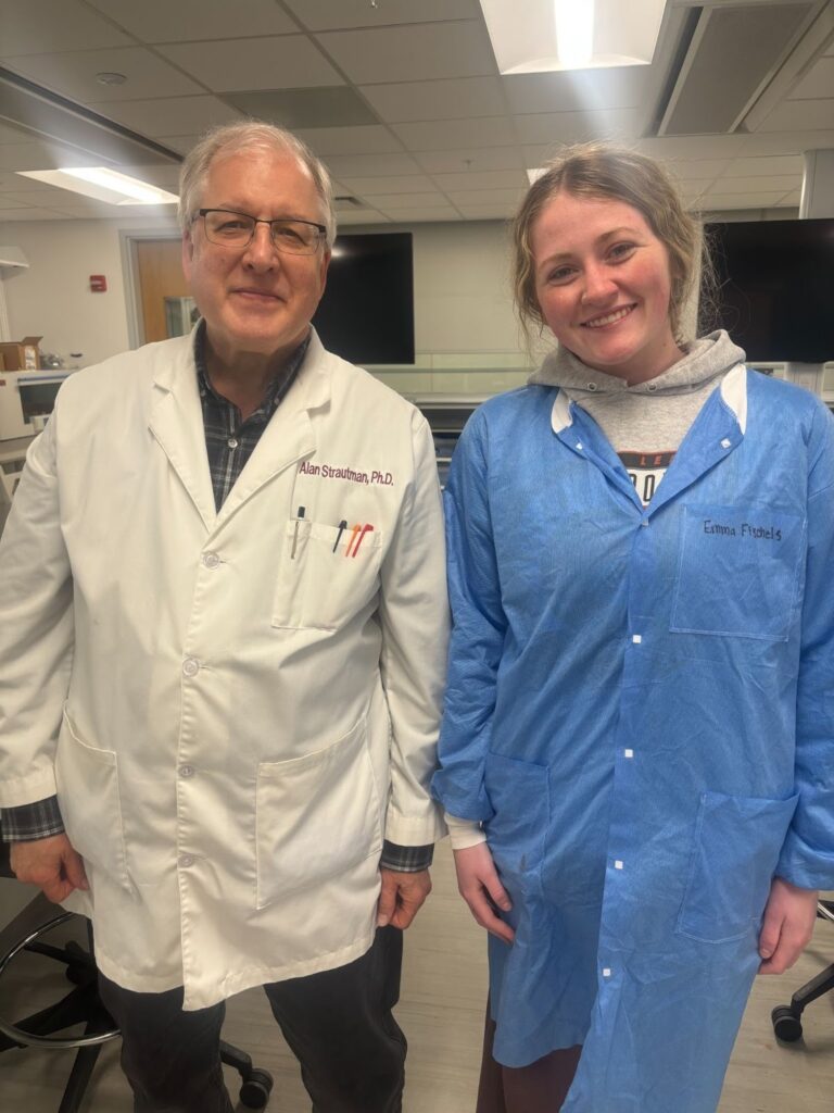 Two researchers stand side by side in a lab: man in a white lab coat with pens in the pocket and glasses, woman in a blue lab coat smiling.