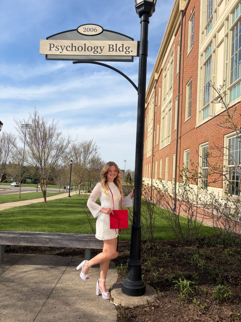 Woman in a white dress carrying a red bag stands beside a black campus street lamp in front of a red brick Psychology Building, on a sunny day (sign reads 'Psychology Bldg.').