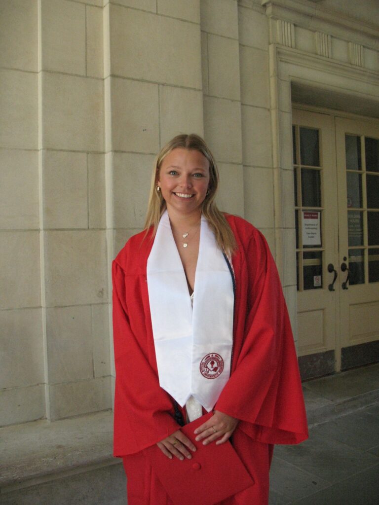 Smiling graduate in a red gown with a white stole, standing outside a beige stone building.