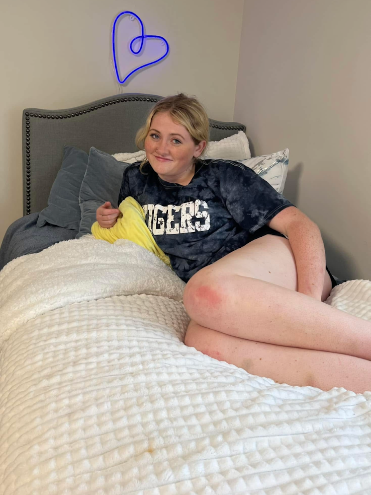 Person sitting up in bed with a blue neon heart wall decoration behind, smiling at the camera.