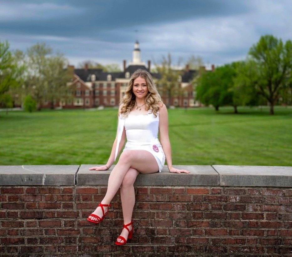 Young woman in a white outfit sits on a brick ledge in a grassy campus with trees and a stately building in the distance under a cloudy sky.