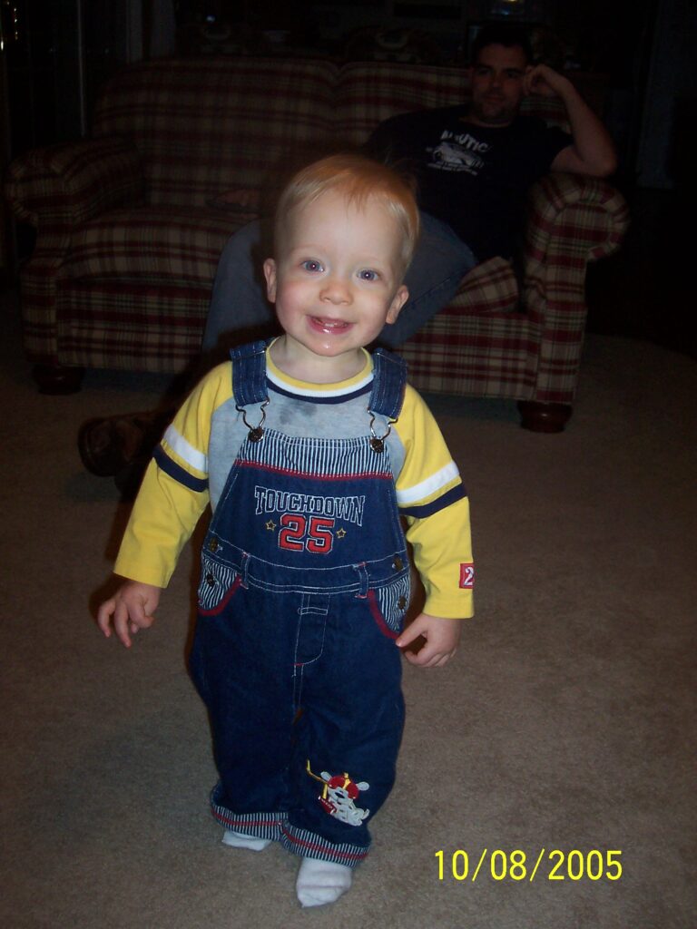 Smiling toddler in a yellow shirt and blue denim overalls standing in a living room; a man sits on a plaid sofa in the background.