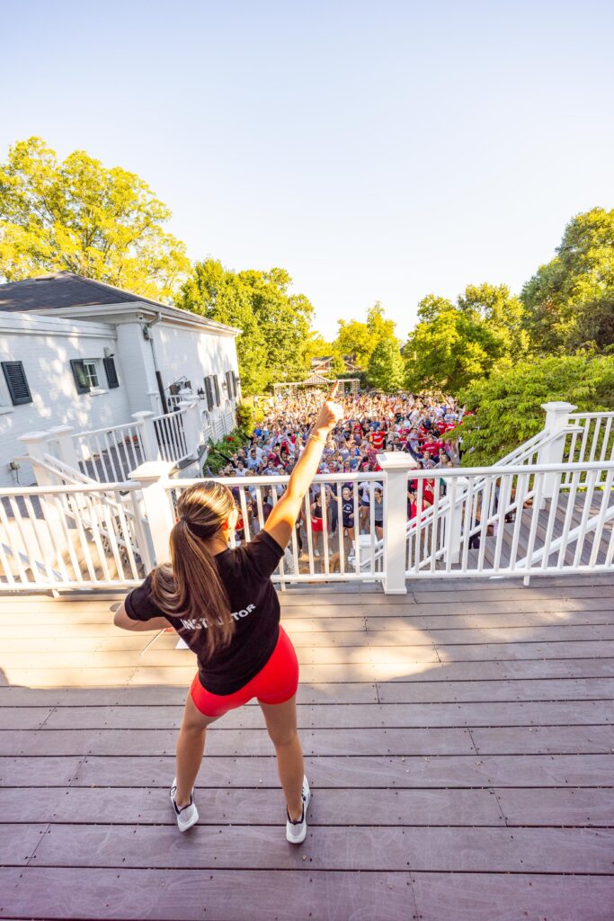 Woman on a wooden deck addressing a large outdoor crowd with her arm raised toward the audience.