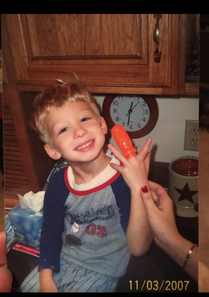 Smiling young boy with blond hair sits in a kitchen, holding an orange toy near his cheek; clock and wooden cabinets in the background.