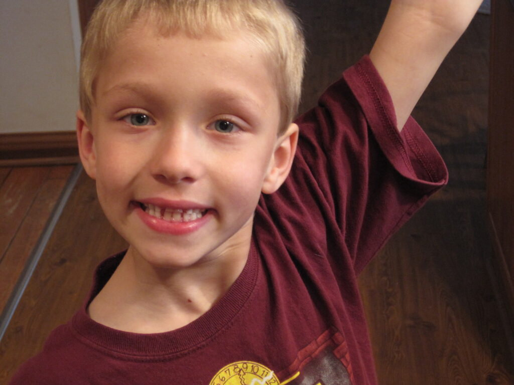 Smiling young boy with light hair and blue eyes, raising an arm in a maroon shirt indoors, camera close to face.