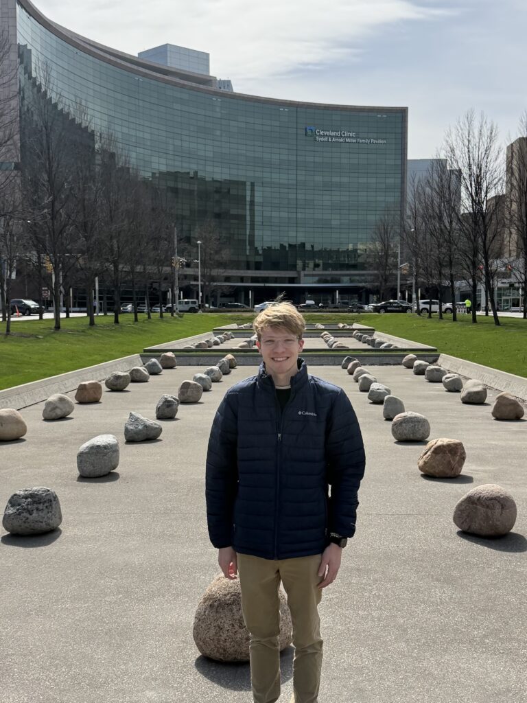 Young man in a dark blue jacket standing on a rock-lined walkway in front of a curved glass Cleveland Clinic building.