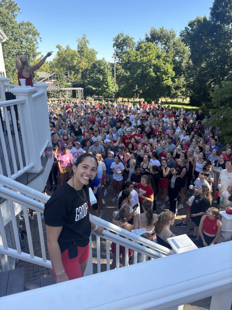 Woman on a white balcony pointing to a large crowd gathered outdoors on a sunny day.