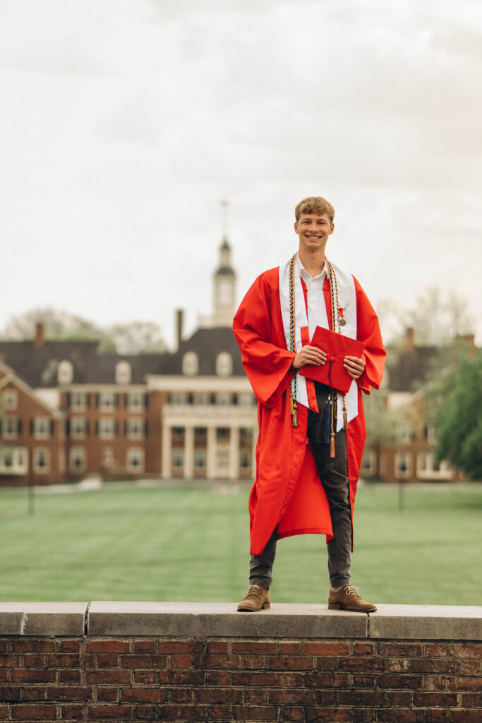 Graduate in a red cap and gown standing on a brick ledge outdoors, smiling at the camera while holding a red mortarboard cap and honor cords around the neck.