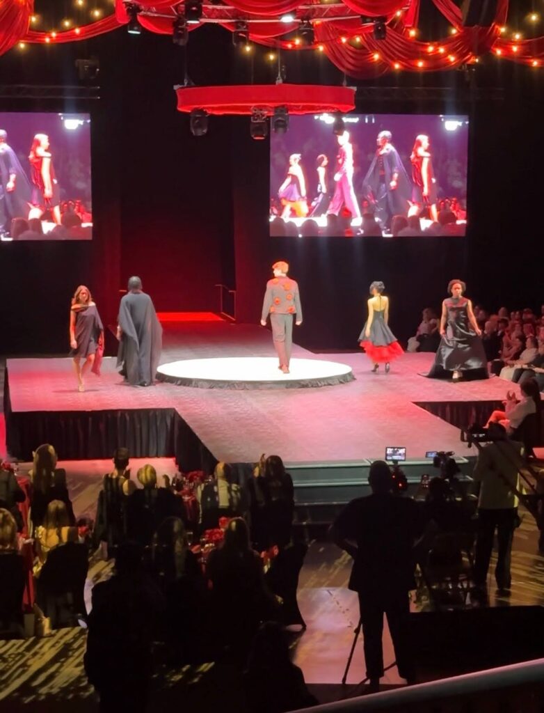 Models walk a circular illuminated runway on a fashion show stage with red drapery overhead and large screens showing the show.