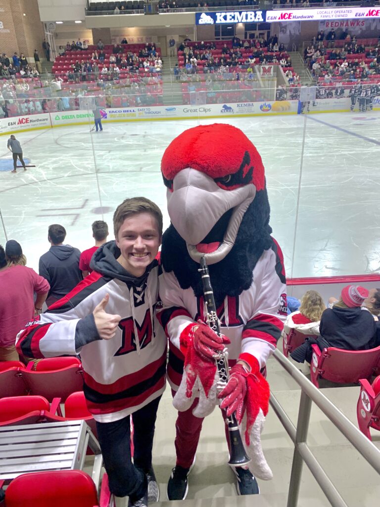 Young man in a red-and-black jacket posing with a red-and-black mascot holding a clarinet at a hockey arena.