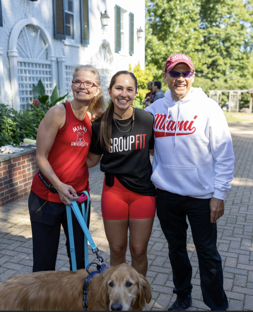 Three people posing together outdoors, wearing Miami-themed athletic apparel, with a golden retriever in the foreground on a leash.
