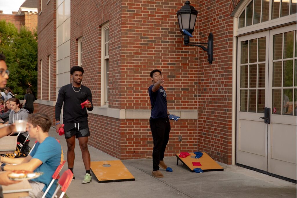 Groups of students outside a brick building, sitting at a table with food and bags, while others stand nearby at a campus event with orange cornhole boards nearby.