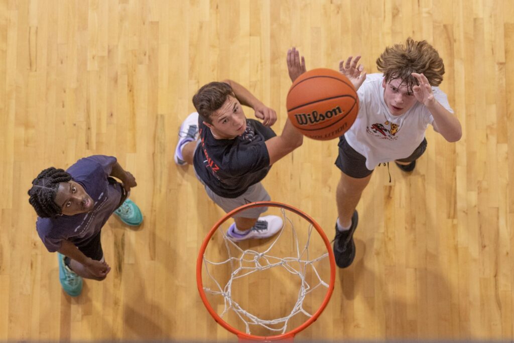 Overhead view of three players jumping to reach a basketball near the hoop on a wooden gym floor.