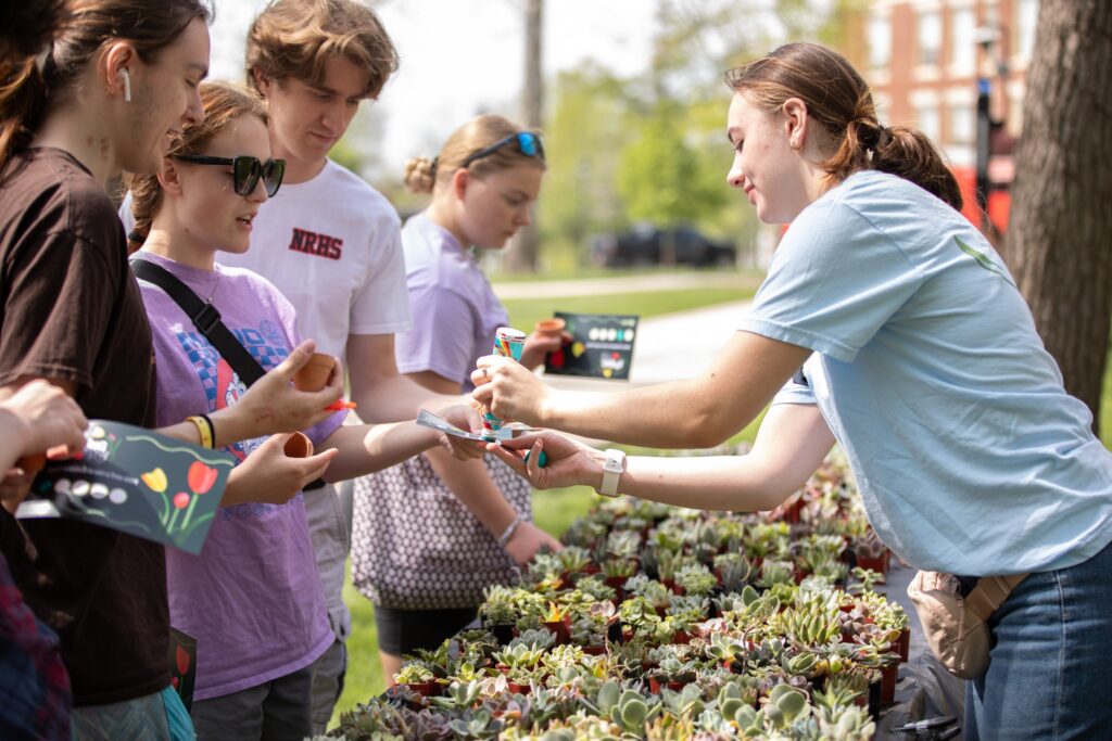 Group of young people at a plant sale exchanging items over rows of succulent pots in bright outdoor light.