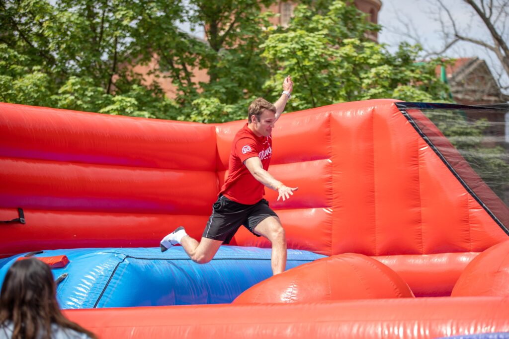 Man in a red shirt hops on a bright orange-red inflatable obstacle course outdoors, raising one arm as he runs forward.