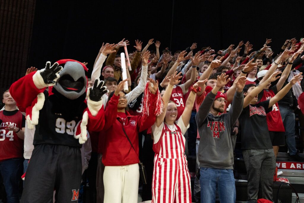 Crowd of Miami University students in red and white cheering with raised hands; mascot in red and black on the left.