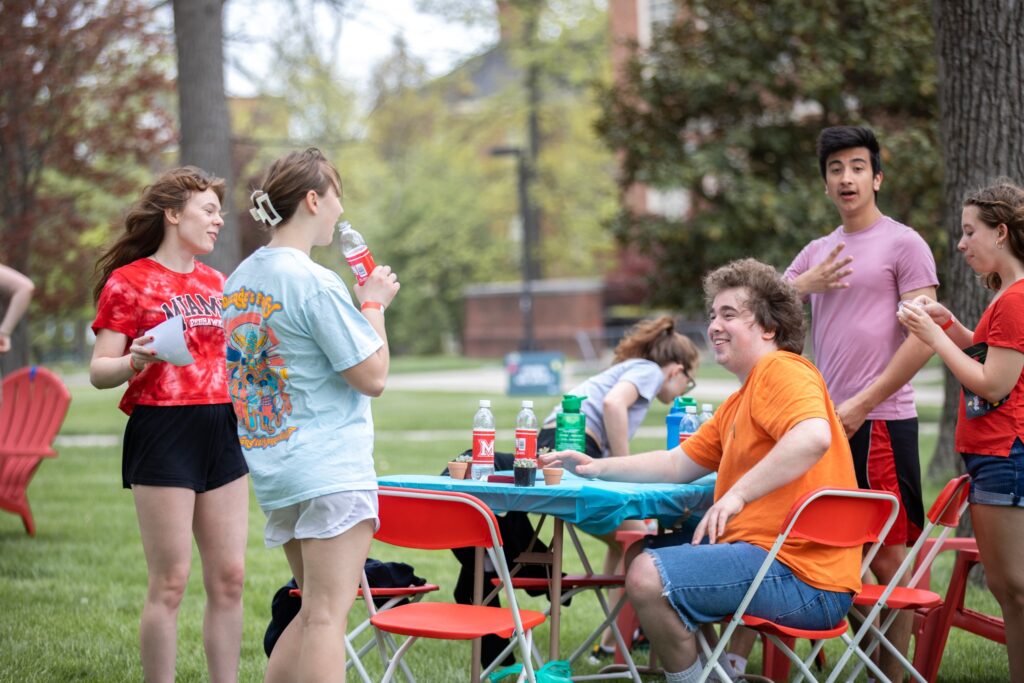 Group of friends hanging out at a park picnic table with drinks and red folding chairs, smiling and chatting.