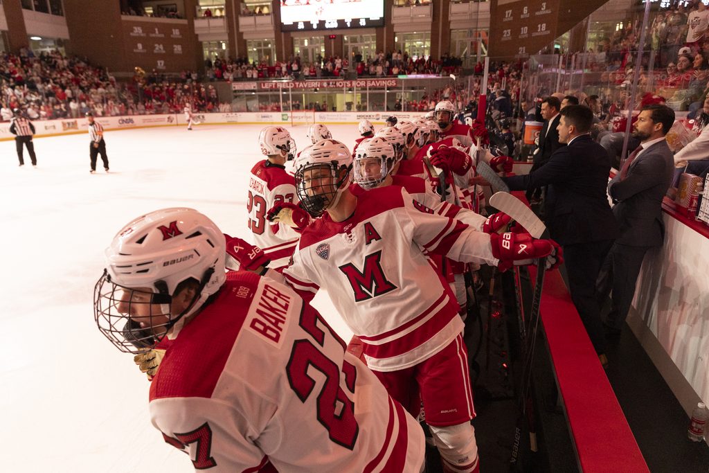 A group of hockey players in red and white uniforms gather near the bench during a game, with spectators and officials visible in the background.