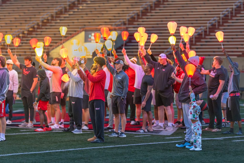 A group of people on a sports field hold up glowing lanterns, standing in formation under evening light.