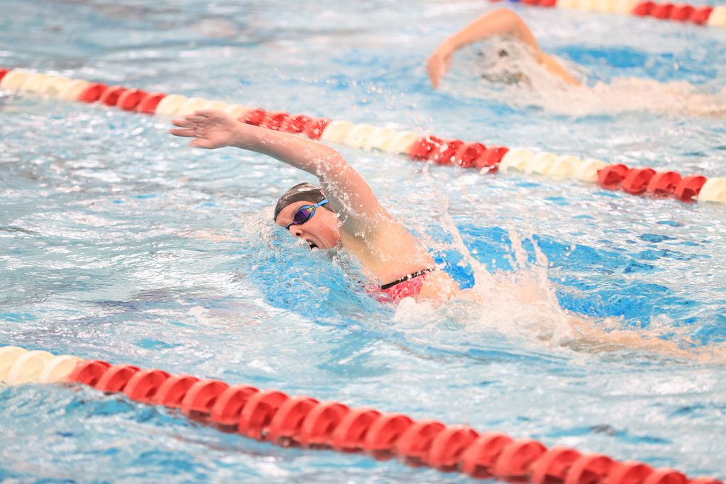 A swimmer in a competitive pool executes a freestyle stroke, with lane dividers visible and another swimmer in the background.