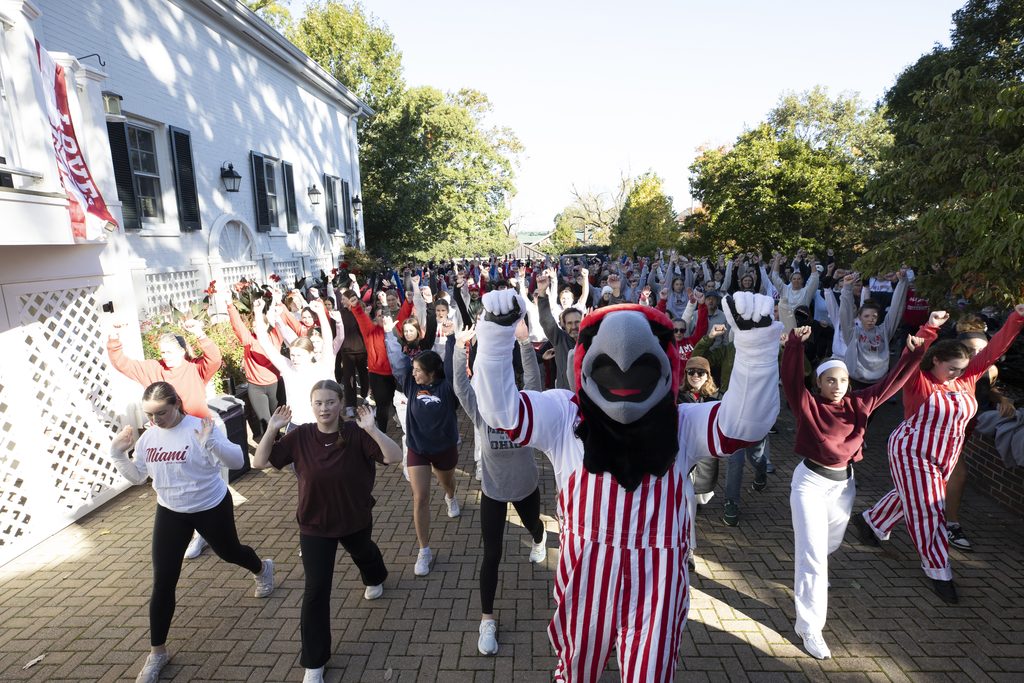 A large group of people, including a mascot in red and white stripes, enthusiastically raise their arms while dancing on a brick patio outside a white building.