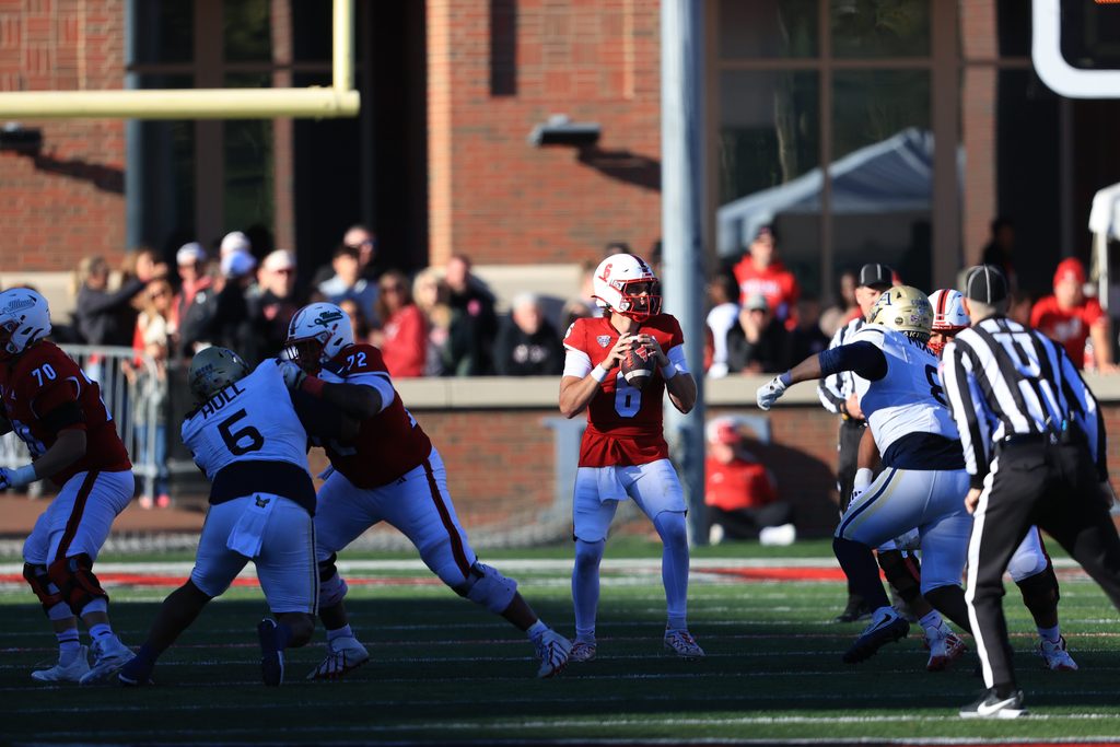 A football quarterback in a red jersey prepares to throw the ball while being surrounded by players on the field during a game.