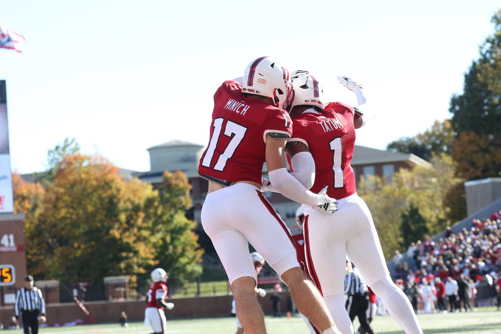Two football players in red uniforms embrace on the field during a game, with spectators and stadium structures visible in the background.